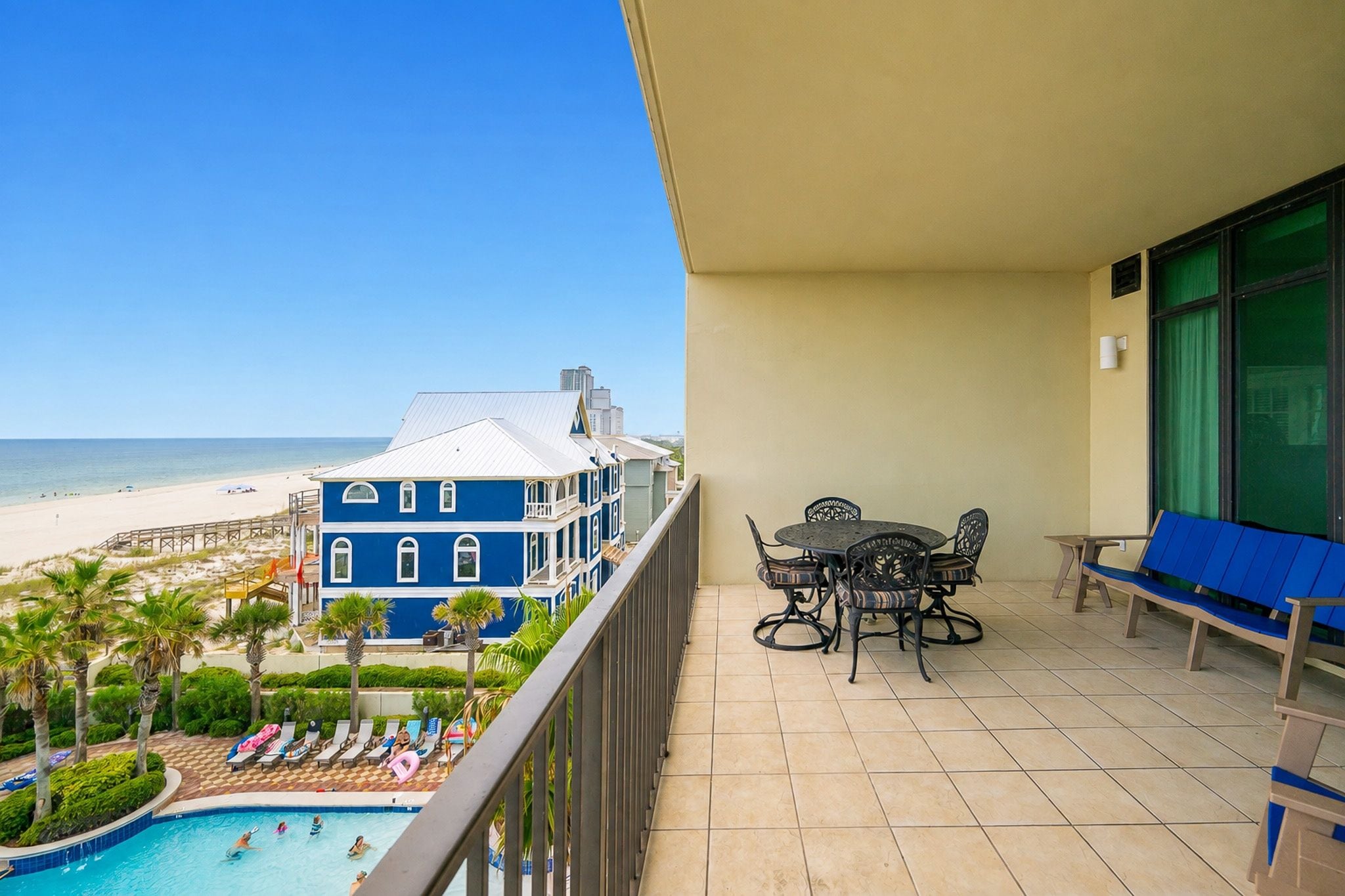 A balcony with a pool, a dining area, and a view of the beach and hotel buildings.