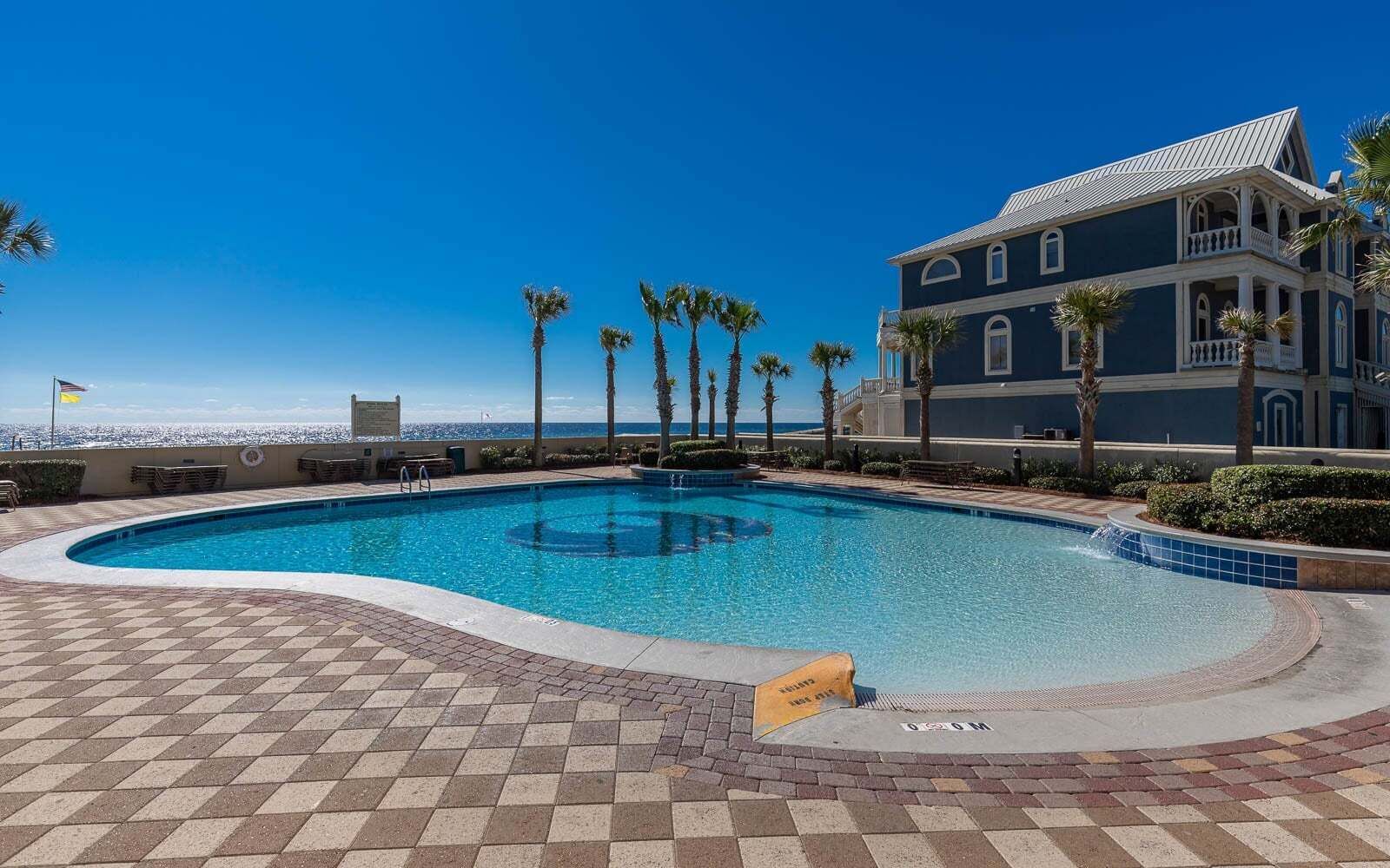 A swimming pool with a tiled deck, palm trees, and a building with a blue facade.
