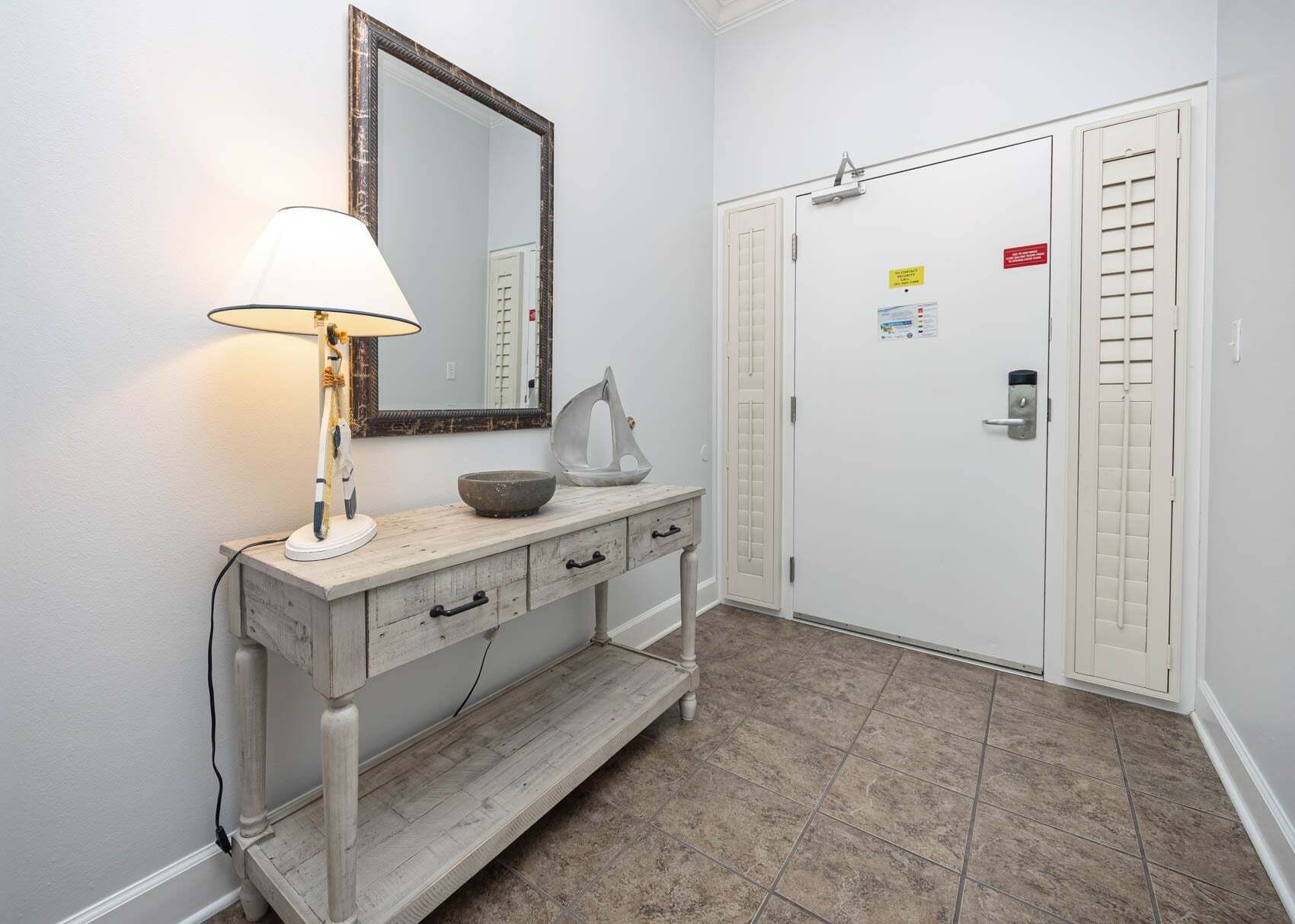 Un couloir avec une porte blanche, un miroir, une lampe, une petite table sur laquelle se trouve un bol, et un sol carrelé.