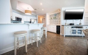 A modern kitchen with white cabinetry, a central island with stools, a built-in oven, and a large flat-screen TV mounted above the island. - Phoenix West 603 3 Bedroom Condo (Orange Beach)
