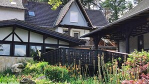 Een traditioneel huis met een puntige dakkapel, een duidelijke veranda en een tuin met diverse planten en bloemen.