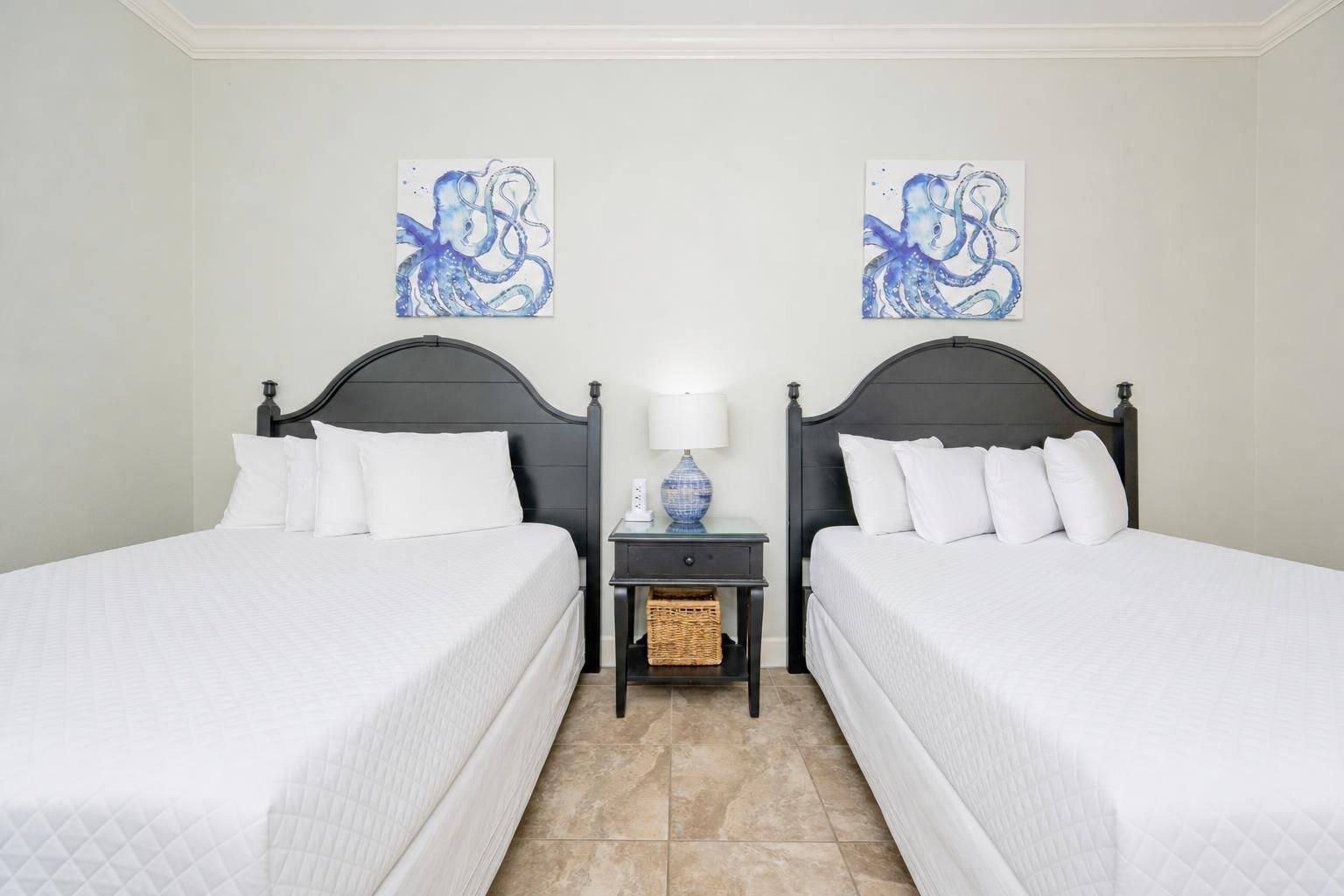 Habitación de hotel con dos camas, una mesita de noche con lámpara y dos obras de arte azules en la pared.