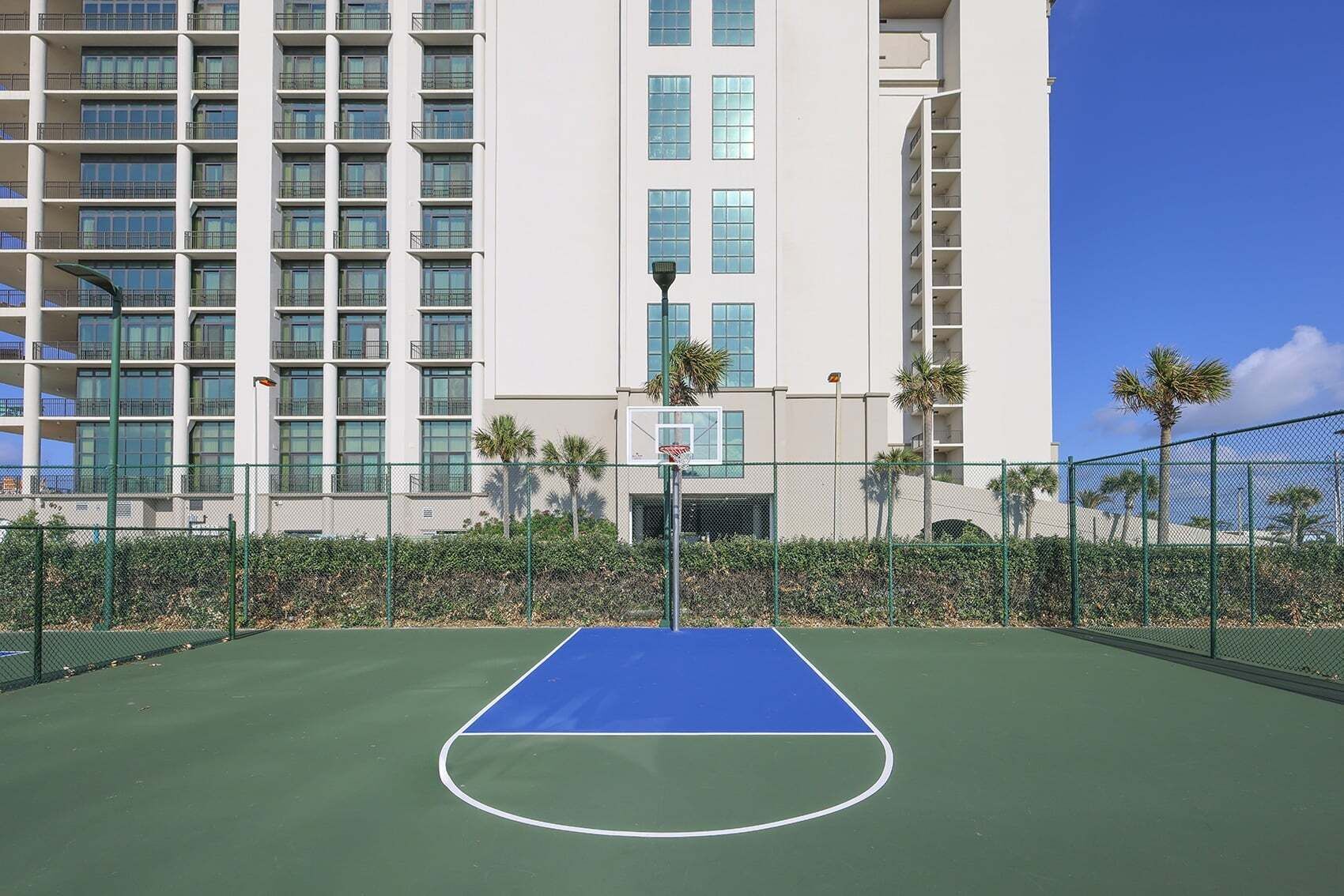 A tennis court with a basketball hoop, surrounded by a fence, and a multi-story building in the background.