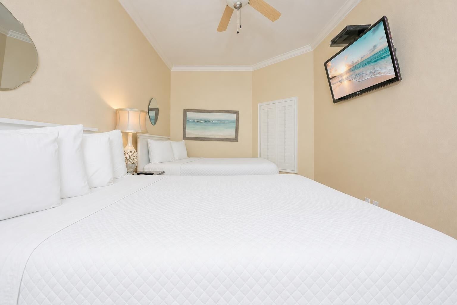 A hotel room with a large bed, a ceiling fan, a wall-mounted TV, and a painting of a beach scene.
