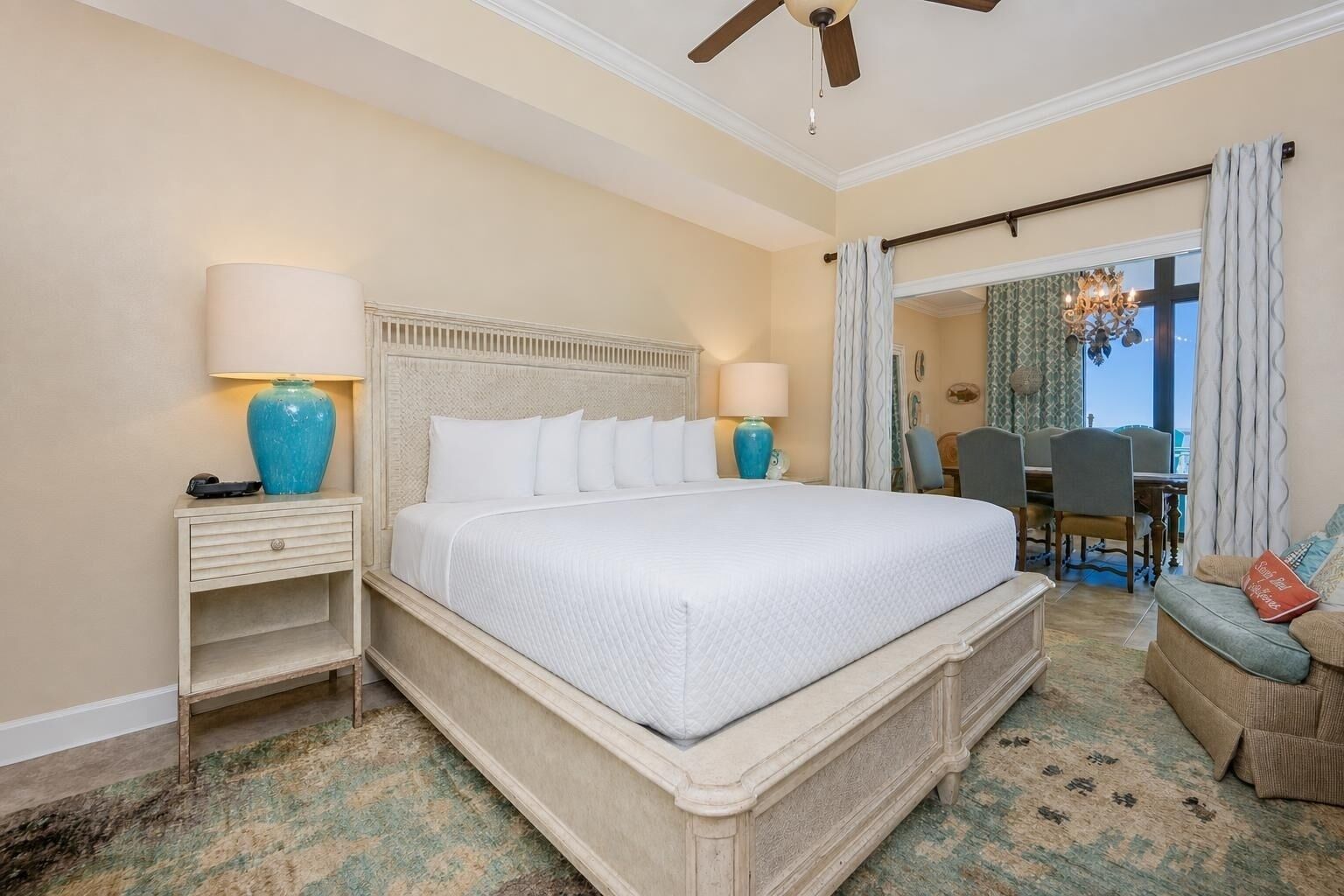 A hotel room with a large bed, two bedside tables, a chandelier, a dining area, and a view of the outdoors.