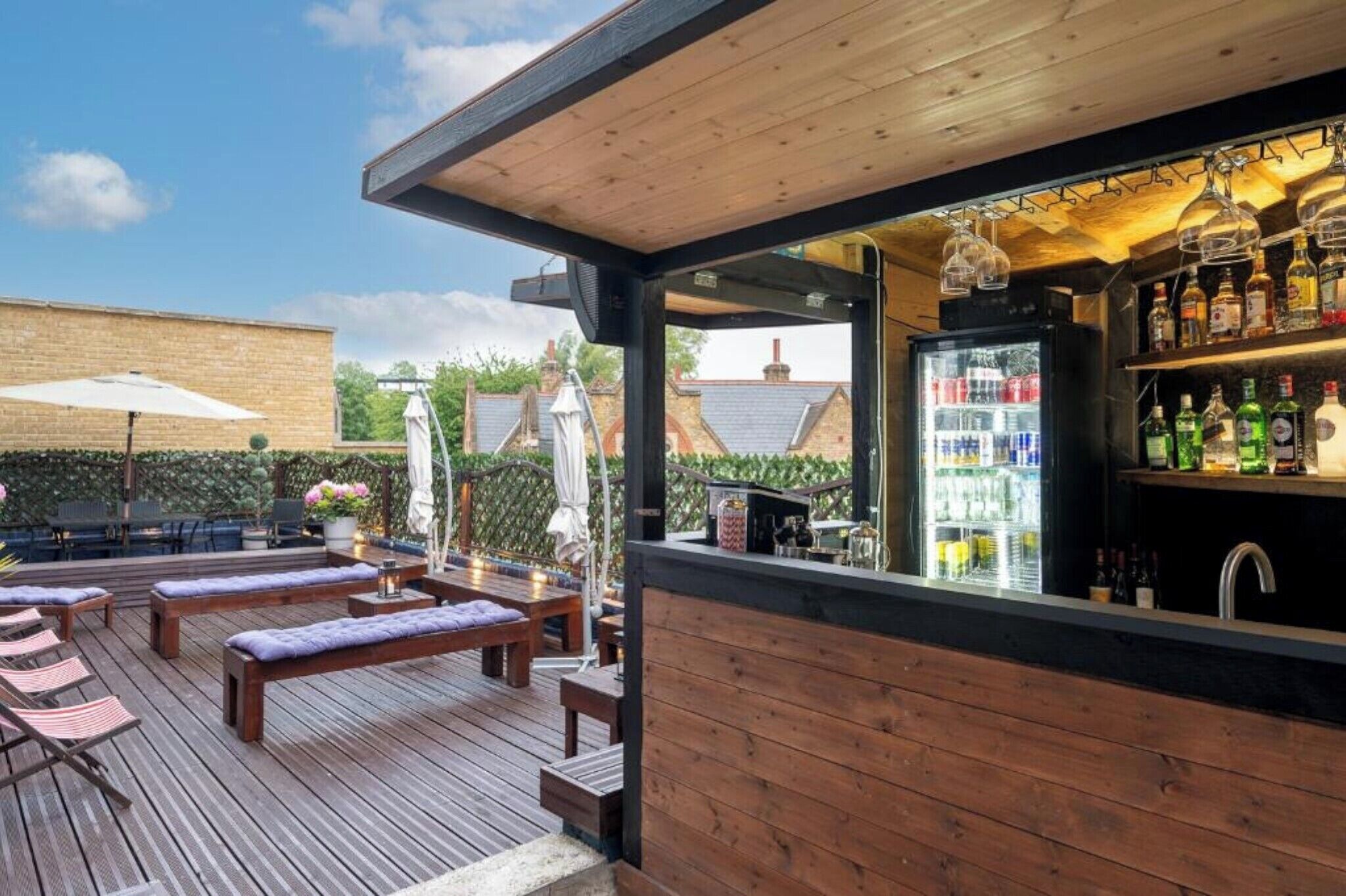 A wooden deck with lounge chairs and a bar area with a refrigerator and shelves of bottles.