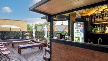 A wooden deck with lounge chairs and a bar area with a refrigerator and shelves of bottles.