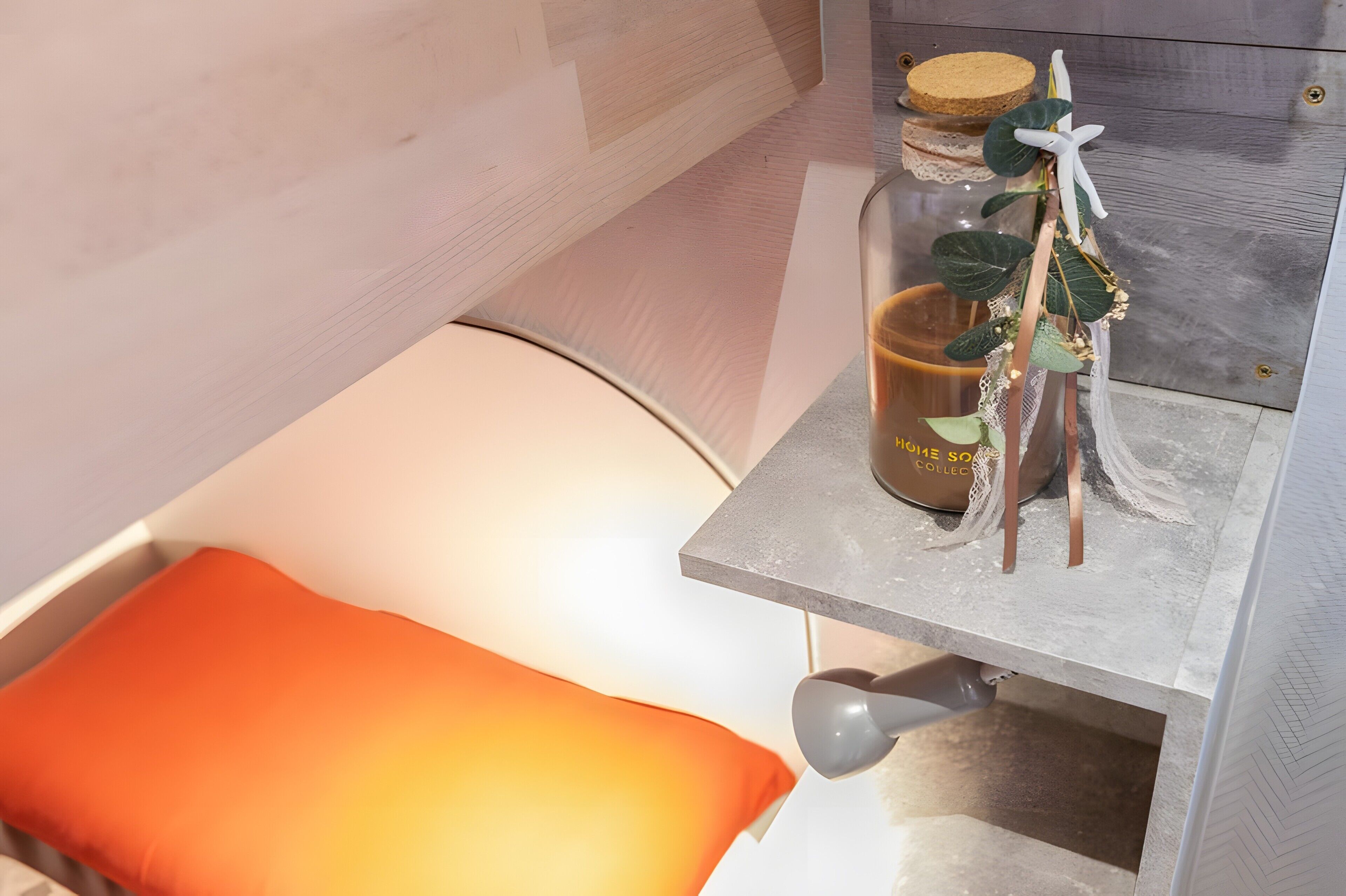 A bed with an orange pillow, a bedside table with a candle, and a small plant.