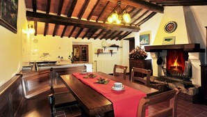 A dining area with a wooden table, chairs, a red tablecloth, a fireplace, and a kitchen in the background. - Poggio alla Rocca (Civitella Paganico)