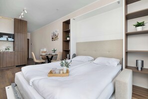 A modern hotel room with a bed, bedside table, a desk with a chair, a small plant, and a wall-mounted shelf. - Air-conditioned Studio with Balcony | Close to Main Station (Kraków)