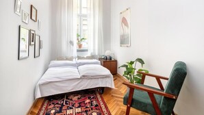 Ein Schlafzimmer mit Bett, Sessel, Fenster mit Vorhängen, einer Wand mit gerahmten Bildern und einer Pendelleuchte.