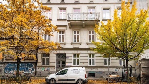 Ein Gebäude mit mehreren Fenstern und Balkonen, ein weißes Auto auf dem Parkplatz und Bäume mit gelben Blättern.