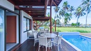 A covered outdoor area with a pool, wicker furniture, and palm trees in the background. - Bangalo no Carneiros Atlantico by CTA (Tamandaré)