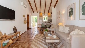 Un grand espace de vie comprenant un canapé, une table basse et un téléviseur. Au fond, on trouve une cuisine avec une table à manger et des chaises.
