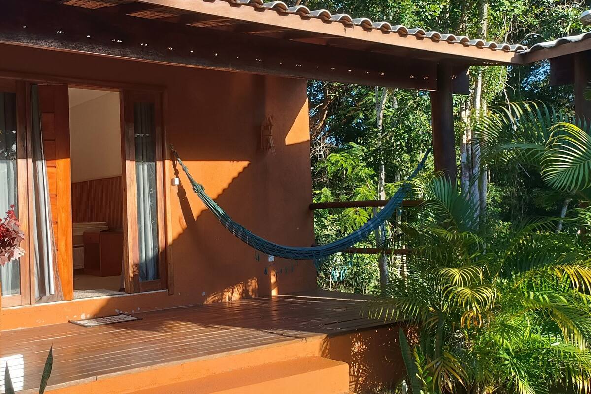 Una terraza de madera con una hamaca, una planta en maceta y vista a una casa con tejado de tejas.