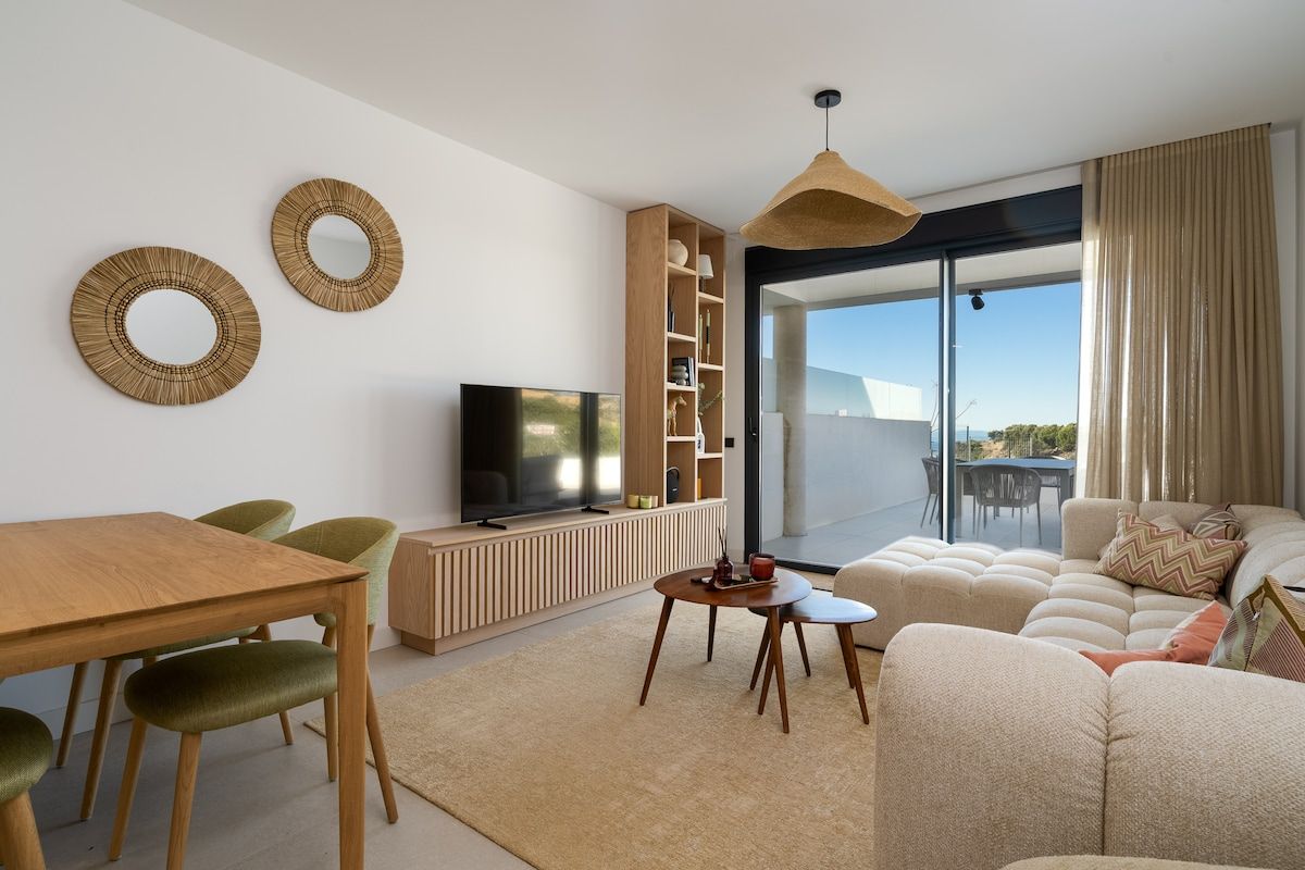 A modern living room with a flat-screen TV, a wooden dining table, green chairs, a beige sofa, and a large window offering a view.