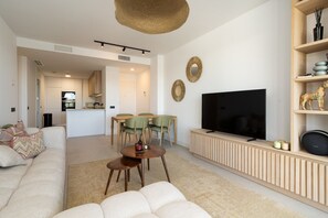 En moderne stue med sofa, fjernsyn, spiseplads med stole og et køkken i baggrunden.