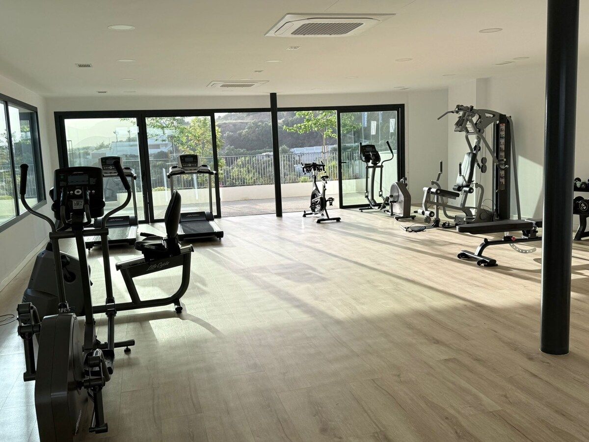 Een fitnessruimte met cardioapparatuur, krachttrainingapparatuur en grote ramen met uitzicht op het omliggende landschap.