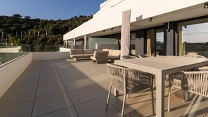 Een modern terras buiten met een eettafel en stoelen, een bank en een glazen balustrade met uitzicht op een beboste heuvel.