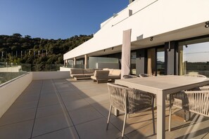 A modern outdoor terrace with a dining table and chairs, a sofa, and a glass balustrade overlooking a forested hillside. - Med Blue by Time & Home (Marbella)