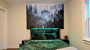 A bedroom with a large forest-themed painting on the wall, a bed with green bedding, and a bedside table with a plant. - ​The Seattle Scenes House: 5-Suite Boutique Home near Light Rail (Seattle)