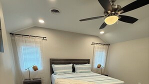 Ein Schlafzimmer mit einem Bett, einem Deckenventilator, zwei Nachttischen, einem Teppich und einem Fenster mit Jalousien.