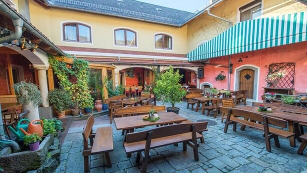 An outdoor patio area with wooden tables and benches, surrounded by potted plants and a stone floor. - Lindengut (Natternbach)