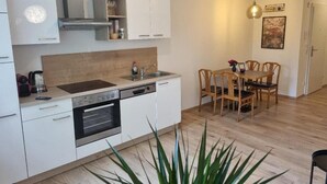 A modern kitchen with white cabinets, a built-in oven, and a dining area with wooden chairs and a table. - Lindengut (Natternbach)