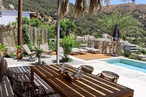 A poolside area with a wooden table and chairs, lounge chairs, and a view of a hillside. - South Palms luxury village house (Rodakino)