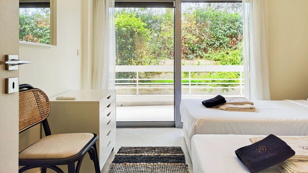 Ein Schlafzimmer mit Bett, Schreibtisch, Stuhl, Klimaanlage und Blick auf Grünanlagen durch eine Schiebetür.