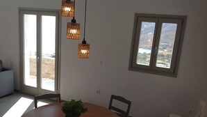 A dining area with a round table, chairs, a potted plant, hanging lights, and a view through a window. - Liopi Village Andros B (Ormos Korthiou)