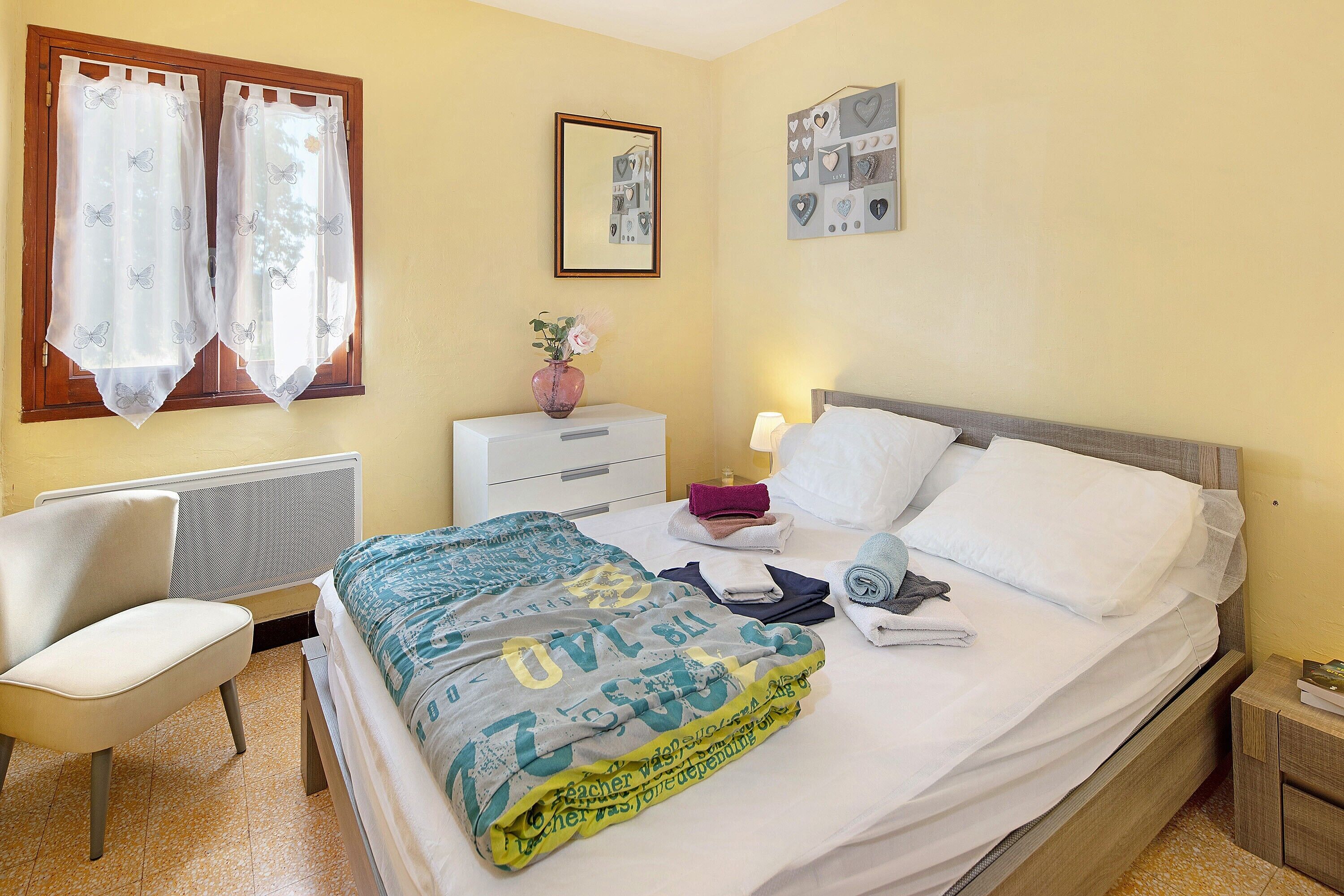 Un lit bien fait, recouvert d’une couverture à motifs, une table de chevet avec une lampe, une chaise et un mur orné d’un tableau encadré.