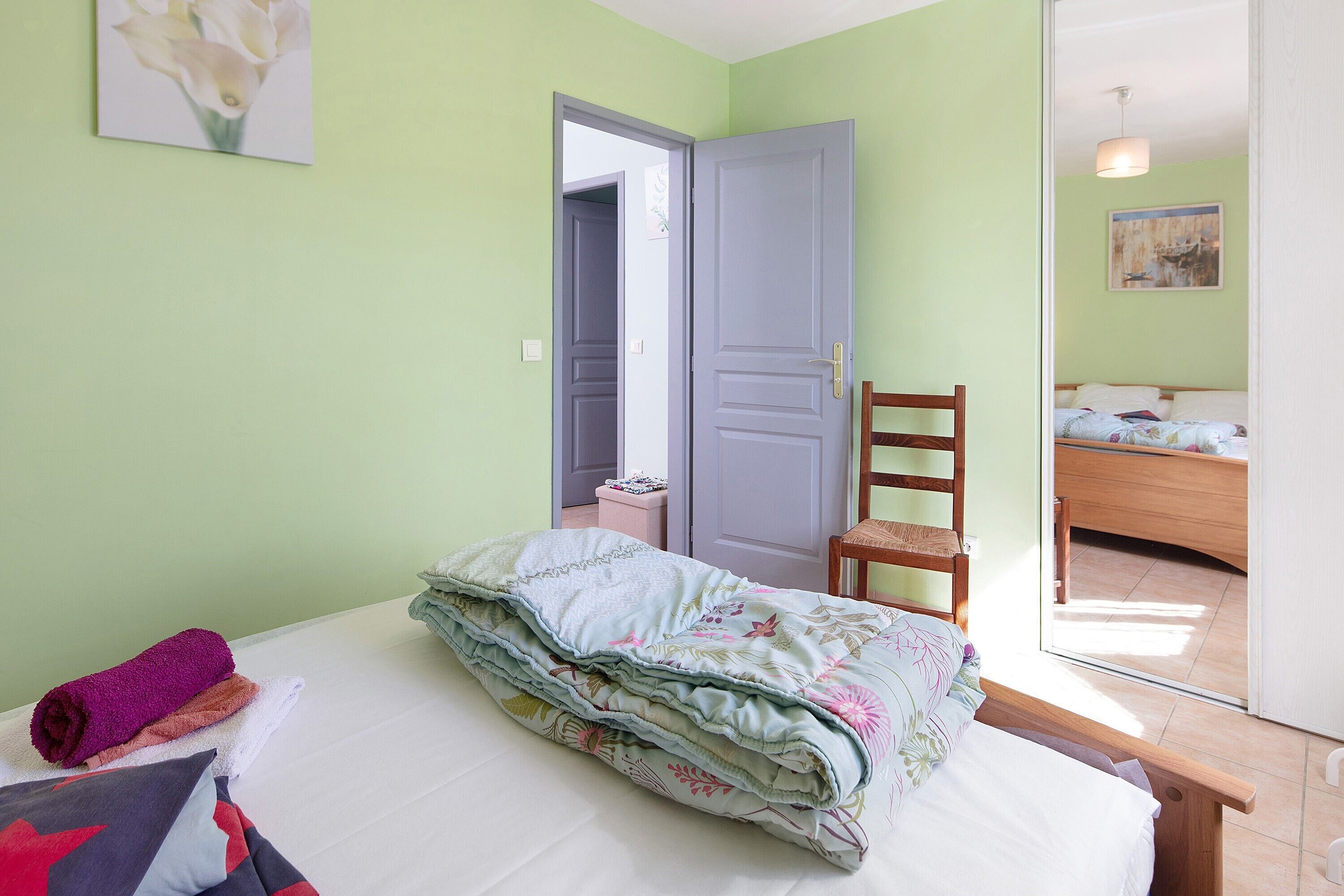 A bedroom with a bed, a chair, a door, and a small table.