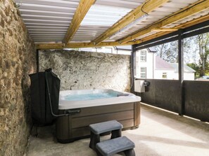 A hot tub in a stone-walled room with a wooden ceiling and a view of a house through a window. - Gate House (Llangefni)