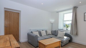 Ein Wohnzimmer mit grauem Sofa, einem Holztisch, einer Holztür und einem Fenster mit Vorhängen.