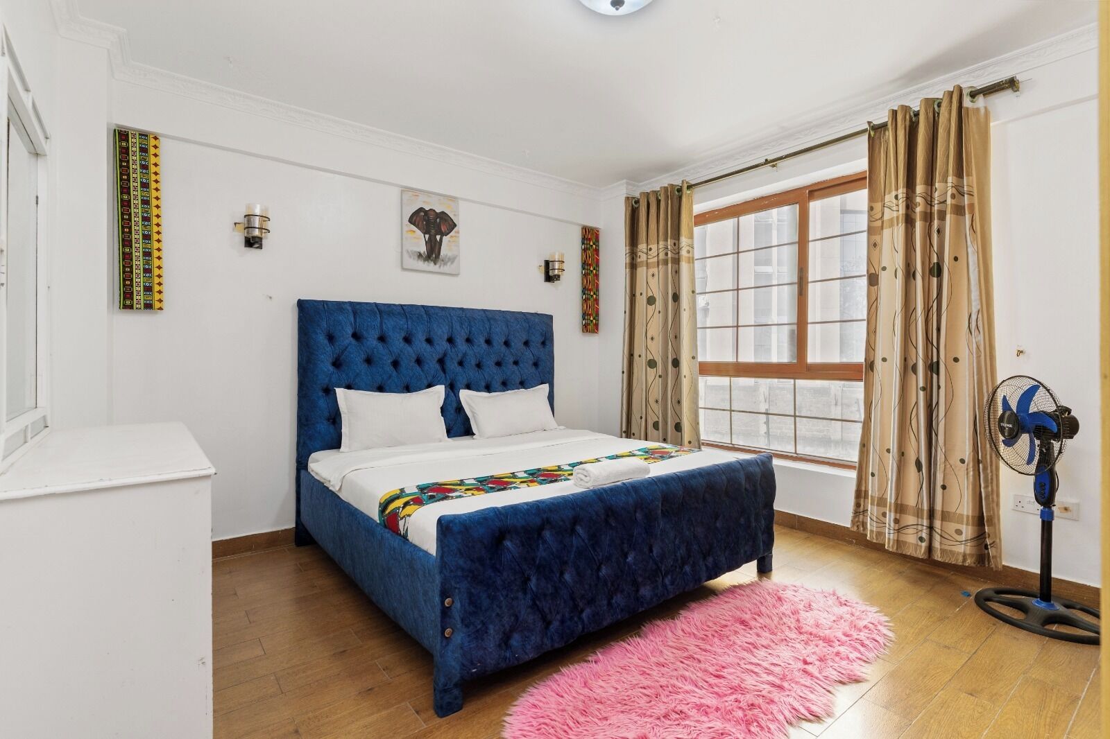 A bedroom with a blue upholstered bed, a pink fluffy rug, a standing fan, and a window with curtains.
