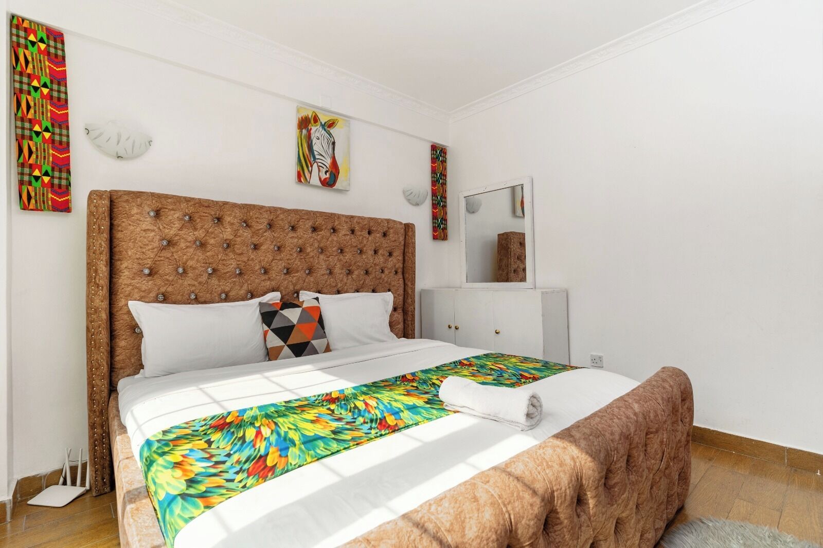 A bedroom with a tufted headboard, a bed with a colorful bedspread, a white bedside table, and a mirror.