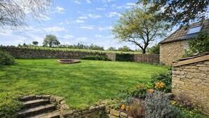 Un jardin entouré de murs en pierre, avec une pelouse, un foyer circulaire et un escalier en pierre menant à un niveau supérieur.