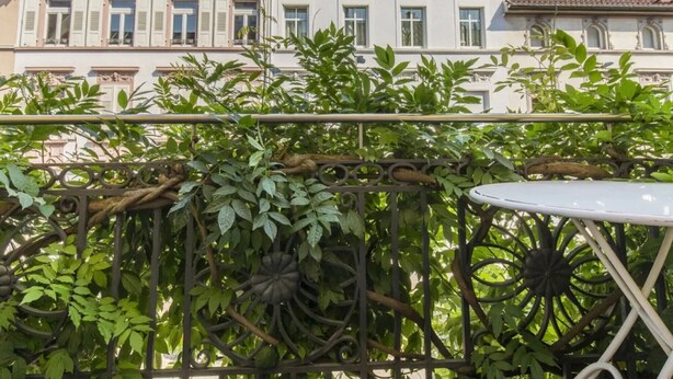 Een balkon met een metalen hekwerk, een witte tafel en weelderige groene beplanting.