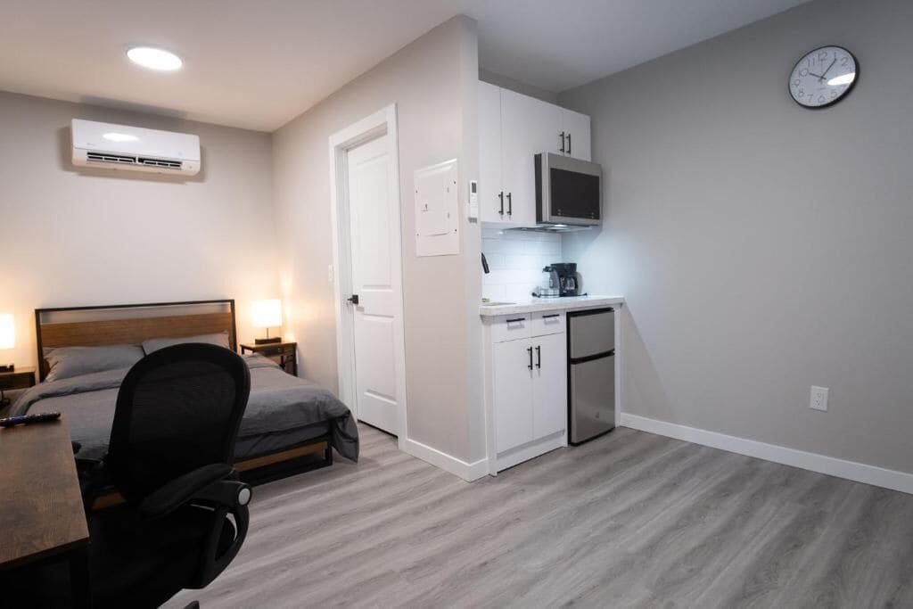 A compact hotel room with a bed, desk, chair, kitchenette, and a clock on the wall.