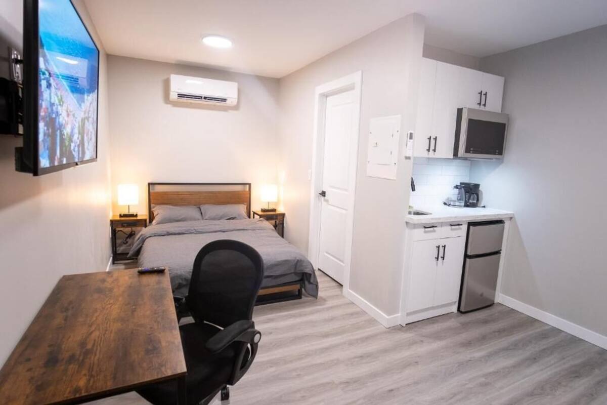 Habitación de hotel pequeña con cama, mesita de noche, silla, escritorio, kitchenette y televisor montado en la pared.