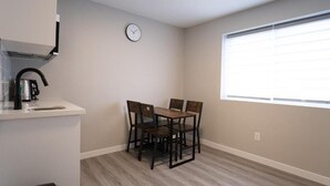 A small kitchenette with a coffee maker, a dining table with four chairs, a clock on the wall, and a window with blinds. - CK Motel (Chatham)