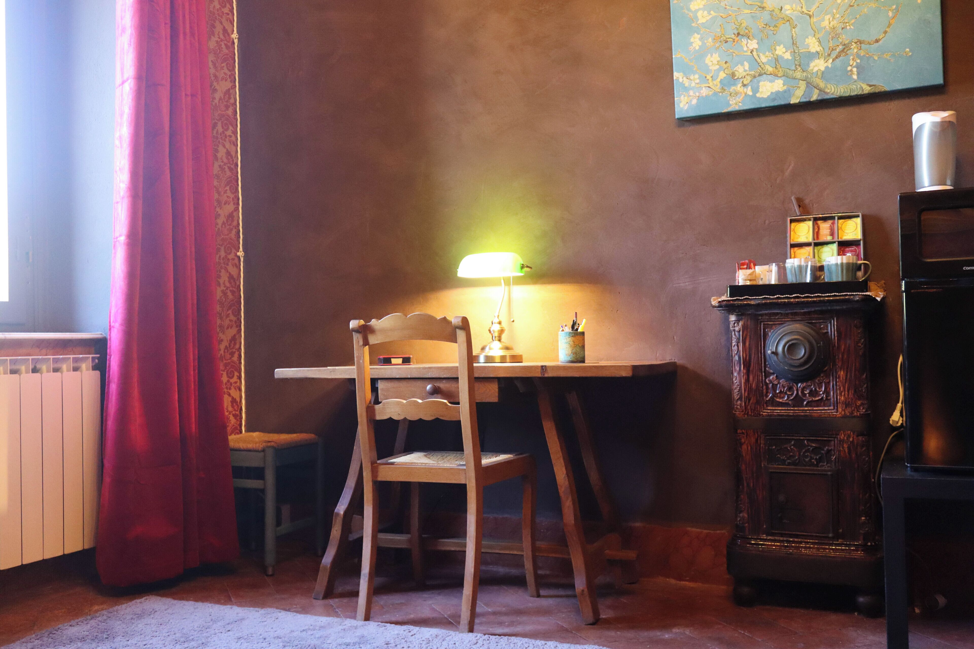 A room with a wooden desk, a chair, a lamp, a radiator, a stove, and a window with curtains.