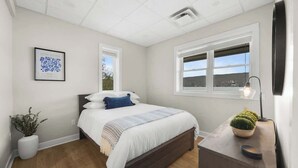 A bedroom with a bed, a bedside table with a lamp, a window, and a framed picture on the wall.