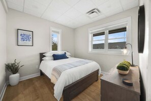A bedroom with a bed, a bedside table with a lamp, a window, and a framed picture on the wall.