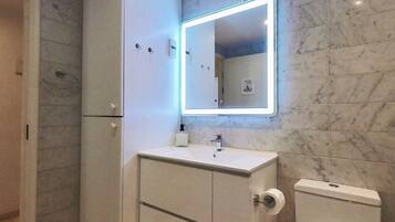 Un baño con pared de mármol, un lavamanos blanco, un espejo con luz y un inodoro.