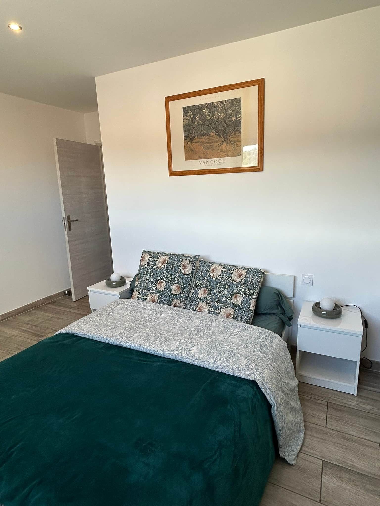 Een slaapkamer met een bed, twee nachtkastjes, een nachttafel, een kunstwerk in een lijst en een deur.