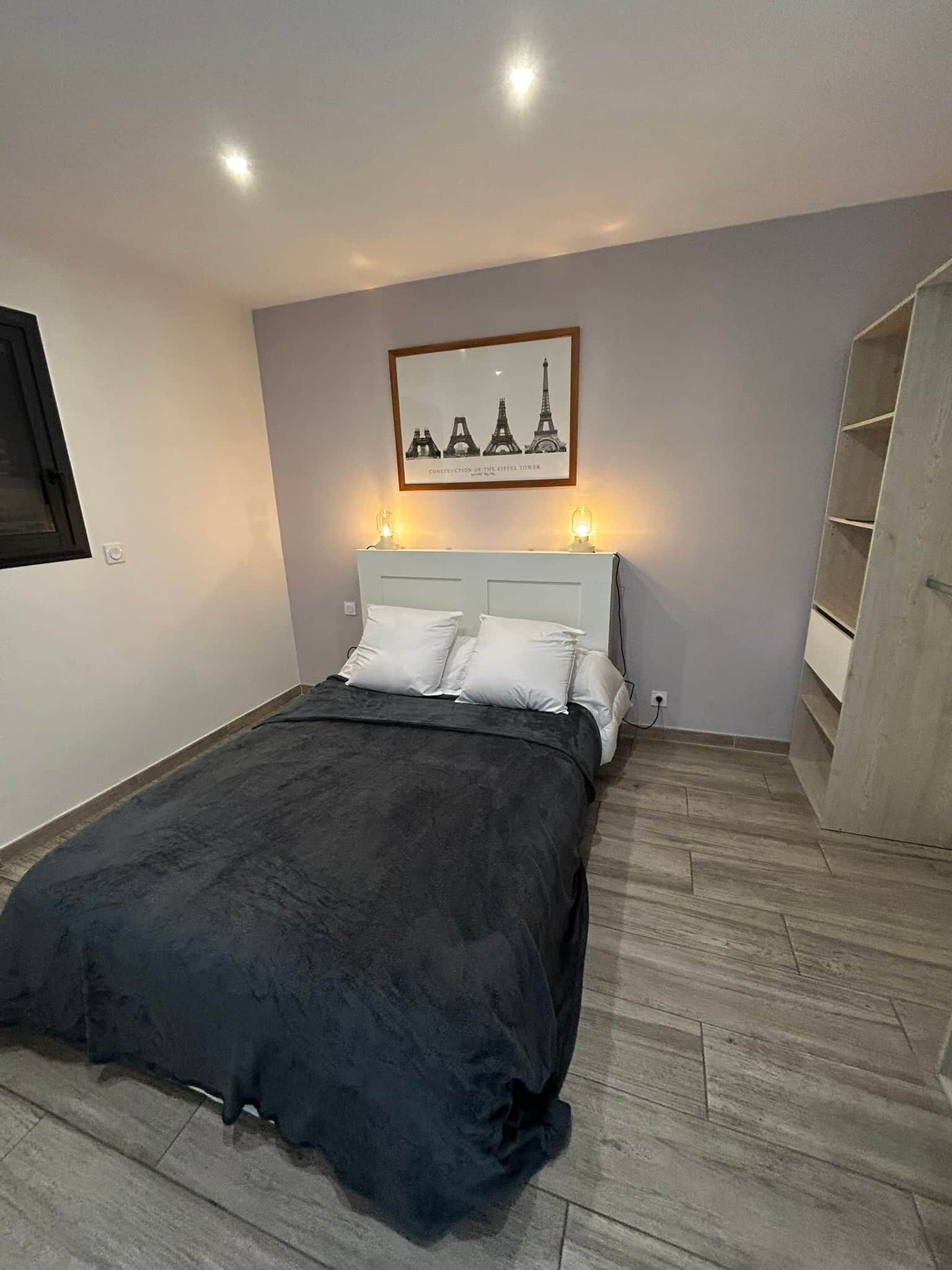 Une chambre à coucher avec un lit, une tête de lit, un tableau représentant la Tour Eiffel accroché au mur et une armoire en bois.
