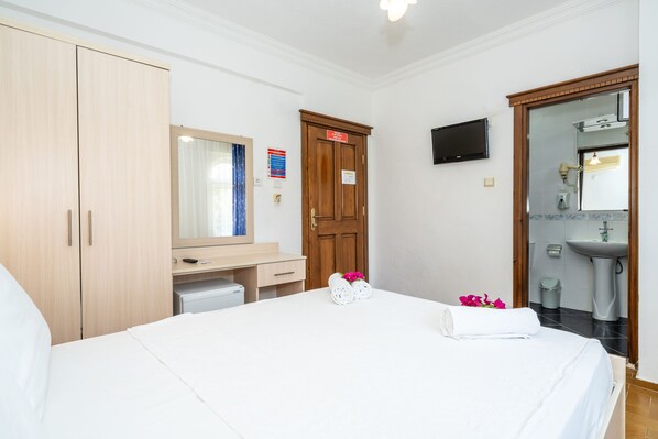 A hotel room with a bed, a wooden wardrobe, a desk with a mirror, a television mounted on the wall, and a bathroom with a sink and mirror. - Gokcen Hotel & Aparments (Fethiye)