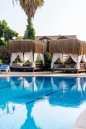 A poolside area with two thatched-roof cabanas, a lounge chair, and a clear blue pool. - Gokcen Hotel & Aparments (Fethiye)